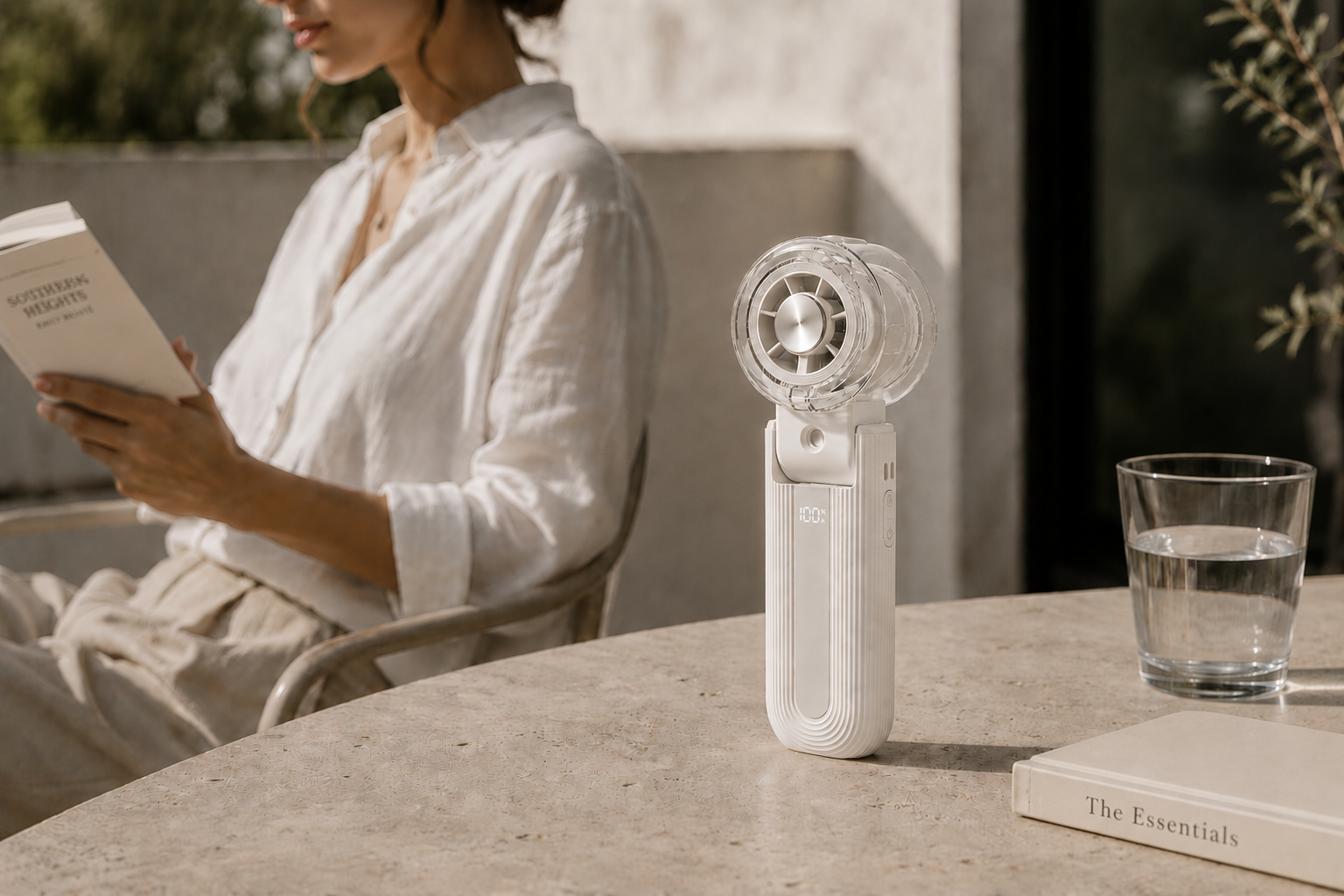 A small white handheld personal fan with a clear circular head stands on a stone table beside a glass of water and a book. In the softly blurred background, a woman in a white blouse sits reading.
The Essentials Edit